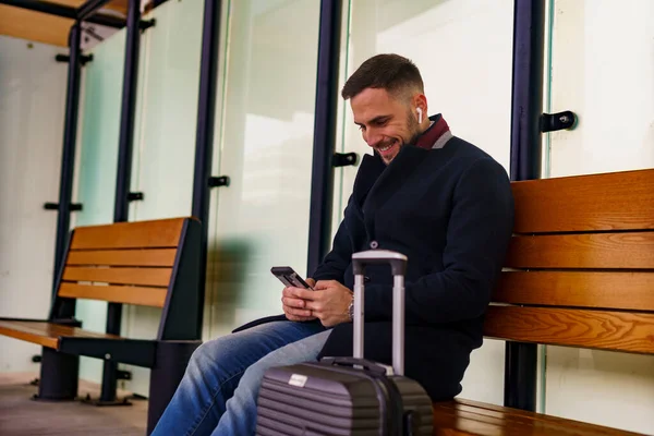 Stock image Young man on a train station smiling at his phone, sitting next his suitcase, he's going on a business trip