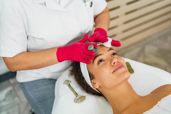 Güzellik uzmanının elleri müşterilerin yüzüne yeşim taşıyla masaj yapıyor. Masaj masasında kaplıcada yatan kadın.