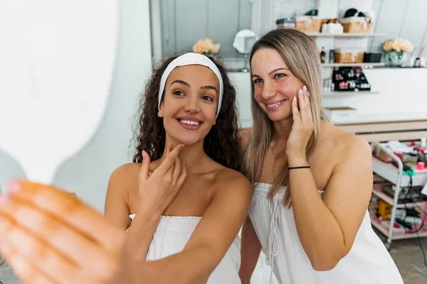 Beyaz havluya sarılı iki genç kadının spa tedavisinden sonra aynada kendilerine baktıkları resim.