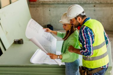 Kadın mimar inşaat alanındaki yöneticiye planı açıklıyor. Mühendis müteahhitle konuşuyor..