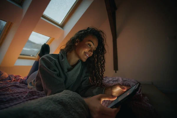 Genç bir kadın uzun bir günün ardından tabletinde film izliyor, gülümsüyor ve eğleniyor.