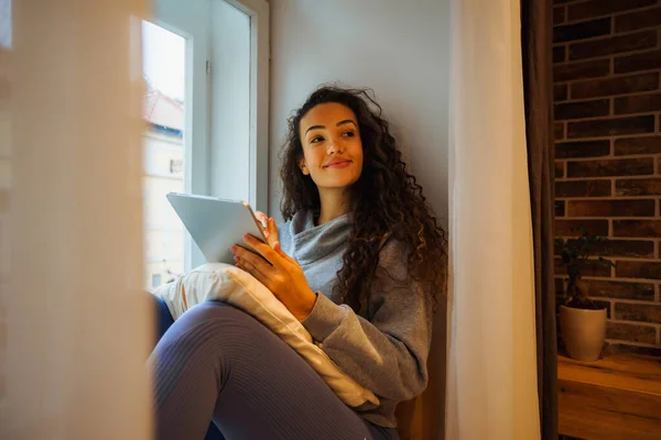 Koyun penceresinde oturan genç bir öğrenci, projelerini düşünüyor ve tabletine planlar yapıyor.