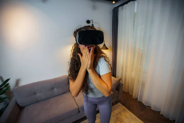 Young woman wearing virtual reality goggles headset, virtual reality box. She\'s astonished at what she sees and covers her mouth with her hands