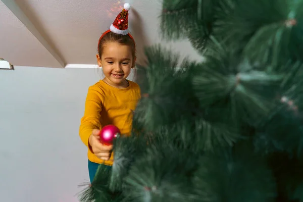 Küçük tatlı kız Noel ağacına pembe bir süs takıyor, şenlikli bir saç bandı takıyor.