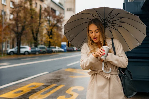 Bulutlu bir gün, sonbaharda yağmurlu bir hava Bir kadın sıcak kahve fincanını tutuyor Toplu taşıma için bekliyor ve saatine bakıyor