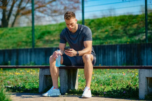 健身后休息 年轻的运动员坐在长椅上 在户外运动后检查手机 — 图库照片