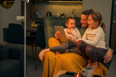Anne ve kızları koltukta rahatça oturup tabletteki videolara bakıyorlar, mutlular ve gülüyorlar.