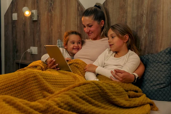 It\'s bedtime and mother is reading a bedtime story to her daughters on her tablet, they are cuddling in bed