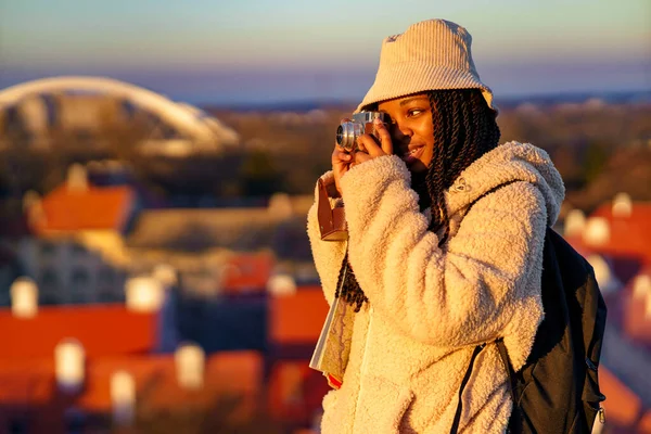 Güzel siyah kadın etrafı geziyor ve arka planda şehir manzarası olan bir fotoğraf makinesiyle konuşuyor.