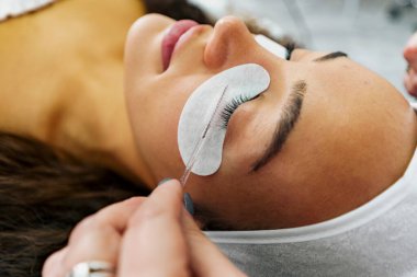 Attractive young woman getting her lashes done at the spa, laying on a massage bed. Beauticians hands doing clients eyelashes
