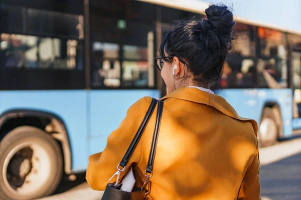 Woman waiting for public transport, headphones in her ears, hair in bun, orange coat with back turned to camera.Bus in the background