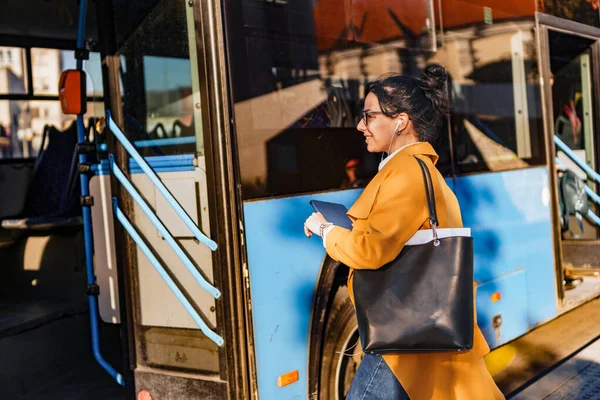 Young woman, student, getting on a bus, bag on her shoulder, hair in a bun, listening to music