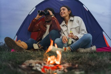 A couple is enjoying themselves in front of a tent by a campfire, the boy is holding binoculars in both hands and watching something in the distance, while the girl is holding two bottles of beverage.