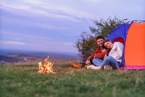 A happy couple is sitting in front of a tent by the camping fire while enjoying the sunset. They enjoy camping in nature.