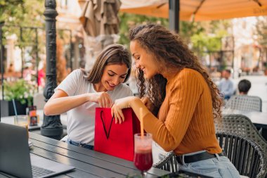 Two girls are sitting in the cafe, pulling out clothes from the shopping bag, and seem thrilled.
