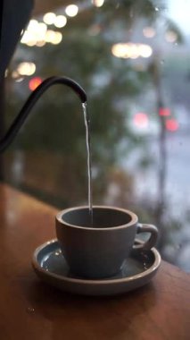 Bardağa sıcak su döken kişi dışarıda yağmur yağarken cam pencerenin yanında kahve yapıyor.