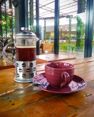 Çay demlenmiş Fransız presi, kahve çekirdekli gümüş bir tutacağa sahip ahşap bir masada oturur. Yanında küçük bir kaşık ile uyumlu bir fincan üzerinde kestane rengi bir seramik bardak var..