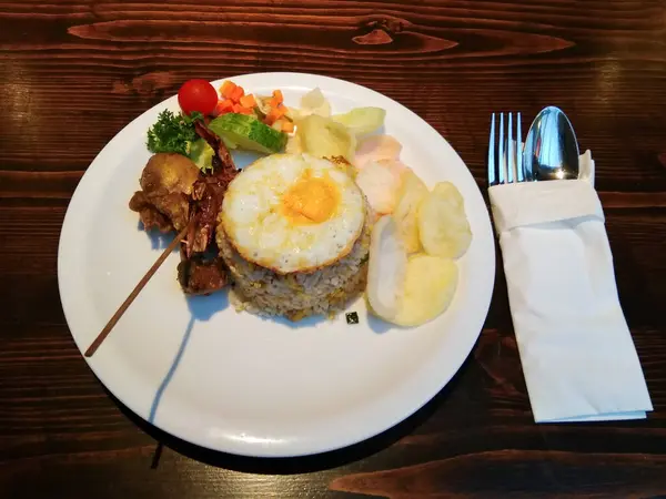 Üzerine sahanda yumurta konmuş pilav beyaz bir tabakta oturur. Pirincin yanında ızgara et ve renkli sebzeler var. Bir karides kraker seçimi doku ekler. 