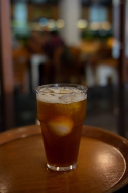 Bir bardak koyu kehribar likörü, muhtemelen buzlu çay, görünür buz küpleriyle, yuvarlak ahşap bir tepside oturur. Arka plan, sıcak ışıklandırmalı bulanık bir iç mekan ayarını gösteriyor