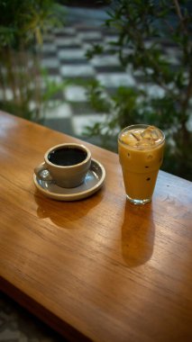 Ahşap bir masa, uyumlu bir tabağın üzerinde gri bir fincan sıcak kahve ve görünür buz küpleriyle birlikte bir bardak buzlu latte tutar. Arka planda bulanık yeşillik görünüyor. Açık hava ya da bahçe ortamını işaret ediyor.