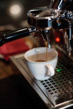 Espresso krom kahve makinesinden beyaz bir porselen fincana akıyor. Makine parlak metal bileşenlerine sahip, yansımaları yakalıyor. Bardak, metal bir damlama tepsisinin üzerine yerleştirilmiş ve delinmiş..