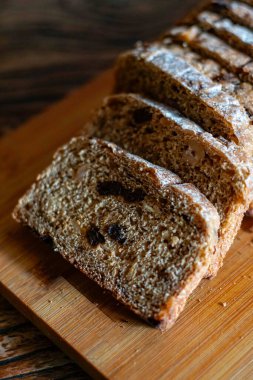 Kuru üzümlü kahverengi ekmek dilimleri tahta kesme tahtasının üzerinde duruyor. Ekmeğin kabuğu hafifçe un ile tozlanmış, ve dokusu yoğun ve sağlıklı görünüyor, kırsal bir görünüm sergiliyor.
