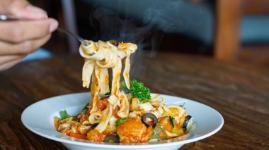 Buharda pişmiş fettucine makarna beyaz bir tabaktan çatalla kaldırılır. Tabakta siyah zeytin, doğranmış domates ve üzerine otlar konmuş ve taze maydanozla süslenmiş kabak yer alıyor. Makarna canlı bir portakal sosuyla kaplıdır.