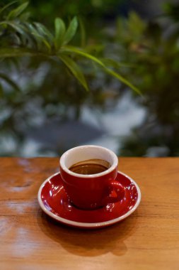 Ahşap masada kırmızı bir fincanda espresso ve bulanık yeşil arka plan. Ayarlar rahat, sıcak bir ortam olduğunu gösteriyor, muhtemelen bir kafe.
