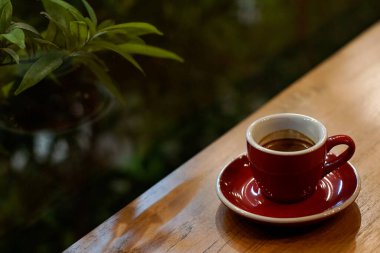 Ahşap masada kırmızı bir fincanda espresso ve bulanık yeşil arka plan. Ayarlar rahat, sıcak bir ortam olduğunu gösteriyor, muhtemelen bir kafe.