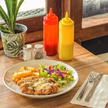 Eritilmiş peynirle kaplanmış ekmek arası tavuk, yanında da patates kızartması ve salata, marul, kırmızı lahana ve domates. Masada tuz ve biber kabı, saksı bitkisi ve kırmızı ve sarı soslar da var.