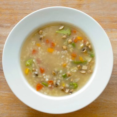 Ahşap bir yüzeye sebze çorbası ve arpa dolu beyaz bir kase yerleştirilir. Çorba hafif ve temiz görünüyor, arpa taneleri etrafa dağıtılmış. Sunum basit, yemeğin kırsal ve sağlıklı doğasını vurguluyor.