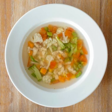 Temiz sebze çorbasıyla dolu beyaz bir seramik kase ahşap bir yüzeye oturur. Çorbada doğranmış portakal havuçları, yeşil brokoli çiçekleri ve beyaz karnabahar parçaları yer alıyor. Genel sunum basit ve sağlıklı bir yemek öneriyor.