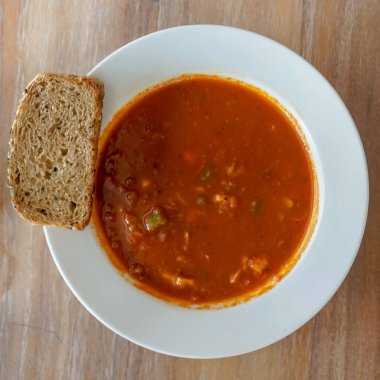 Beyaz seramik tabakta domates çorbası var. Rengi bol, sebze parçaları var. Bu da sağlıklı bir dokuya işaret ediyor. Tabağın kenarında bir dilim çoklu tahıllı ekmek var. Kaba bir doku ve görünür tohumlar var..