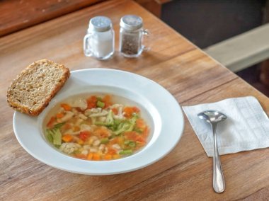 Brokoli, havuç ve dolma biber içeren temiz sebze çorbasıyla dolu beyaz tabak ahşap bir masada oturur. Tabağın kenarında bir dilim çoklu tahıllı ekmek duruyor. Yakınlarda tuzluk ve biberlik ve kaşık beyaz bir peçeteye konur..