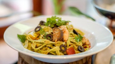 Beyaz bir tabakta servis edilen renkli makarna, üzerinde siyah zeytin dilimleri, sarı dolma biber ve taze maydanoz bulunan tagliatelle..