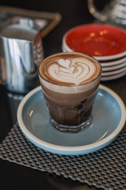 Kalp tasarımı içeren karışık latte sanatıyla kaplı bir bardak katmanlı kahve içeceği. Açık mavi bir tabağın üzerinde, berrak bir bardakta servis edilir, desenli bir tabağa dayanır.
