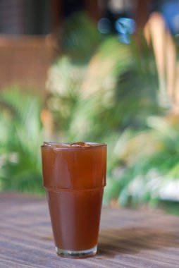 Bir bardak koyu kahverengi sıvı, muhtemelen çay ya da kola, ahşap bir masada oturur. Arkaplan özellikleri bulanık yeşil yeşillik, açık hava ayarlarını gösterir.