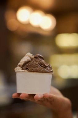 Bir avuç çikolata ve bej dondurmayla dolu beyaz bir kağıt kaseyi tutan bir el. Arka plan bulanık, dondurmayı renklendiriyor.. 