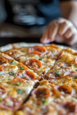 Biberli pizza dilimlenmiş, pepperoni dilimleri ve taze fesleğenle kaplı, ahşap servis tahtasının üzerinde dinleniyor. Peynir erimiş ve hafifçe altın renginde. Bir el pizzaya uzanıyor, sıradan bir yemek düzenini işaret ediyor.