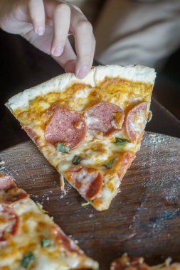 Bir çocuğun eli bir dilim pepperonili pizza tutuyor. Pizzada biraz kızarmış, erimiş peynirli ve üzerinde bir dilim pepperoni olan kıtır bir kabuk var.