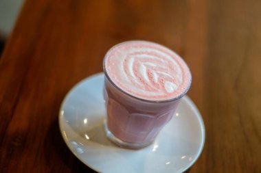 Bir bardak kırmızı kadife latte ahşap bir masanın üzerinde beyaz bir fincan tabağı üzerine konur. Lattelerin yüzeyi, kırmızı ve beyaz kıvrımlı karmaşık köpük sanatı sergiliyor. Görsel olarak çekici bir desen oluşturuyor..