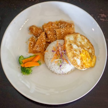 Beyaz tabakta, üzerinde doğranmış mor lahana ve havuçlar olan bir pirinç yığını var. Kızarmış yumurta ve susam tohumuyla kaplanmış kızarmış tavuk parçalarının yanında. Tabağa buğulanmış havuç çubukları ve brokoli çiçeği eşlik ediyor..