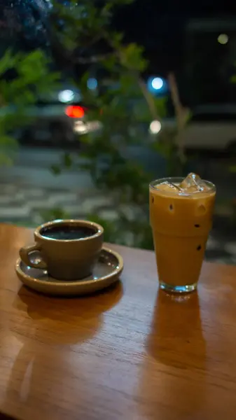 Ahşap bir masa, uyumlu bir tabağın üzerinde gri bir fincan sıcak kahve ve görünür buz küpleriyle birlikte bir bardak buzlu latte tutar. Arka planda bulanık yeşillik görünüyor. Açık hava ya da bahçe ortamını işaret ediyor.