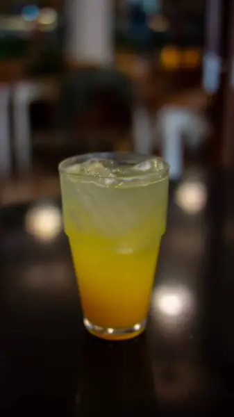 Yarısı portakal sarısı sıvıyla dolu, üstü buz küpleriyle kaplı bir bardak. Meyve suyu, alttaki turuncudan üstteki açık sarıya doğru olan bir gradyanı gösteriyor. Cam koyu, yansıtıcı bir yüzeye yerleştirilmiş.