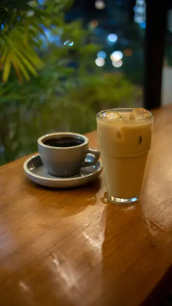 Ahşap bir masa, uyumlu bir tabağın üzerinde gri bir fincan sıcak kahve ve görünür buz küpleriyle birlikte bir bardak buzlu latte tutar. Arka planda bulanık yeşillik görünüyor. Açık hava ya da bahçe ortamını işaret ediyor.