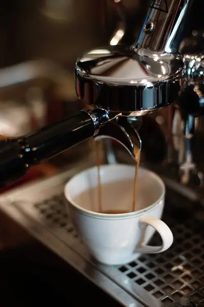 Espresso krom kahve makinesinden beyaz bir porselen fincana akıyor. Makine parlak metal bileşenlerine sahip, yansımaları yakalıyor. Bardak, metal bir damlama tepsisinin üzerine yerleştirilmiş ve delinmiş..