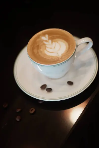 Beyaz seramik bir fincan, bir yaprak desenini andıran karışık köpüklü bir latte içeren uyumlu bir fincan üzerinde oturur. Kurulum koyu bir masanın üzerinde duruyor, yanında dağınık kahve çekirdekleri var, estetiği güçlendiriyor.
