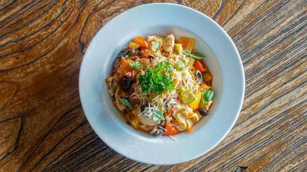 Pasta primavera beyaz bir kasede servis edilir, tahta bir yüzey üzerinde dinlenir. Tabakta maydanoz ve rendelenmiş peynirle süslenmiş çan biberi, siyah zeytin ve brokoli de dahil olmak üzere sebze karışımı yer alıyor..