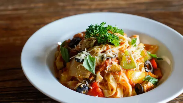 Domates sosu, rendelenmiş peynir, dilimlenmiş siyah zeytin, doğranmış dolma biber ve fesleğen yapraklarıyla kaplı beyaz bir kasede servis edilen makarna tabağı. Taze maydanozla süslenmiş renkli malzemeler iştah açıcı bir sunum yaratıyor.