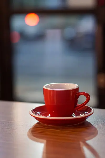 Ahşap masada kırmızı bir fincanda espresso ve bulanık bir trafik geçmişi var. Ayarlar rahat, sıcak bir ortam olduğunu gösteriyor, muhtemelen bir kafe.
